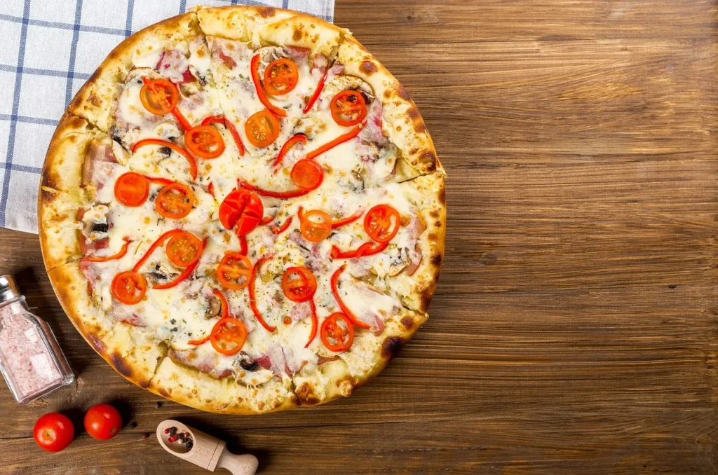 Pizza z pomidorkami koktajlowymi i papryką na drewnianym blacie. Obok pomidory i przyprawy oraz ścierka w kratkę