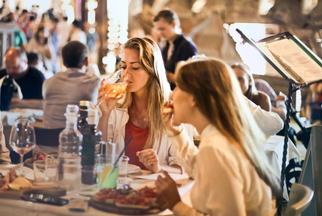 Restauracja pełna gości. Na pierwszym tle kobieta jedząca pizzę i kobieta pijąca drinka, obie siedzą przy jednym stole